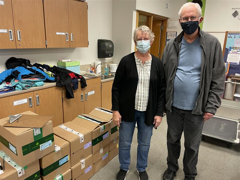 A photo of two grown ups, wearing medical masks, standing by cardboard boxes containing food.