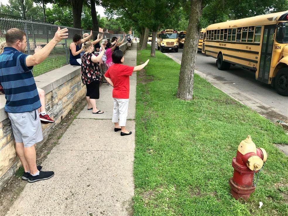 A group of adults waves at school buses as they pull away from a curb.
